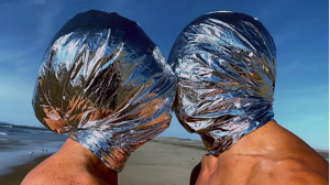 Image of artwork by Ana Teresa Fernandez, The Space Between Us, 2022. Two people with foil wrapped around their heads lean in for a kiss with blue sky in the background.