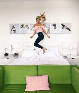 Blonde woman with ponytail wearing a pink shirt and pants jumps on a white bed with a green sofa in the foreground and photos of birds on the wall behind her