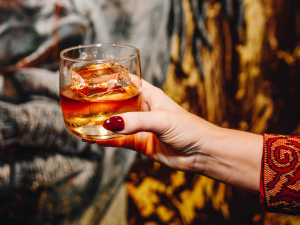 Close up image of a manicured hand holding a glass of bourbon with a piece of art in the background