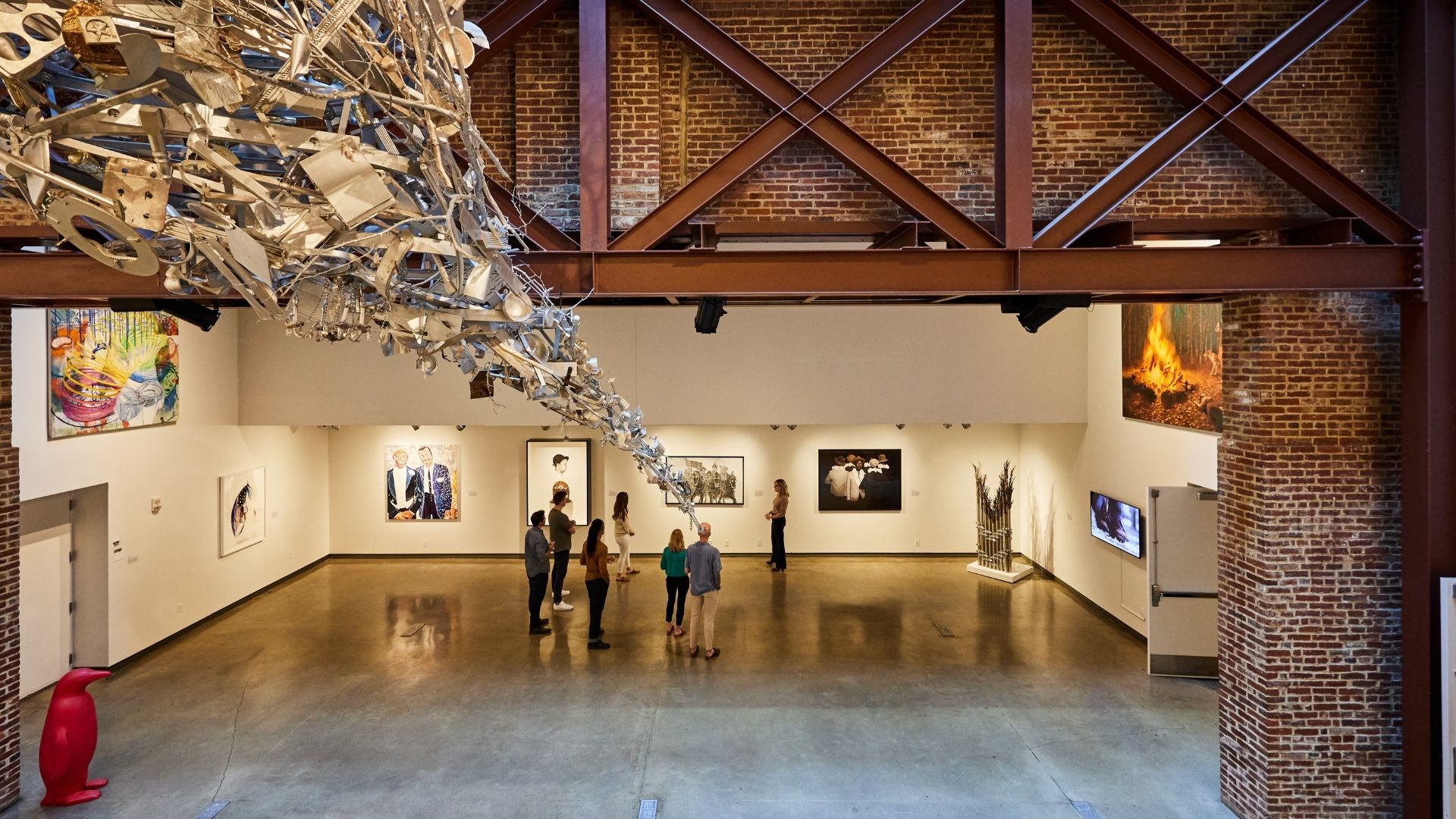 Large gallery space with metal structure hanging from the ceiling and people looking at paintings on the wall