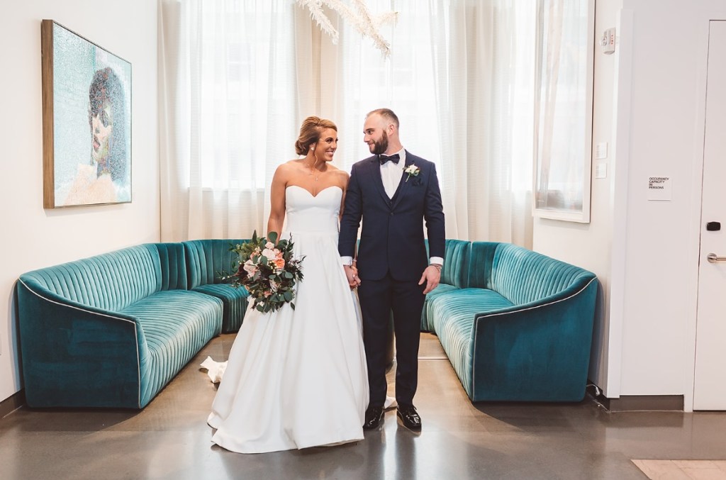 Bride and Groom hold hands and stand in front of a large turquoise sectional