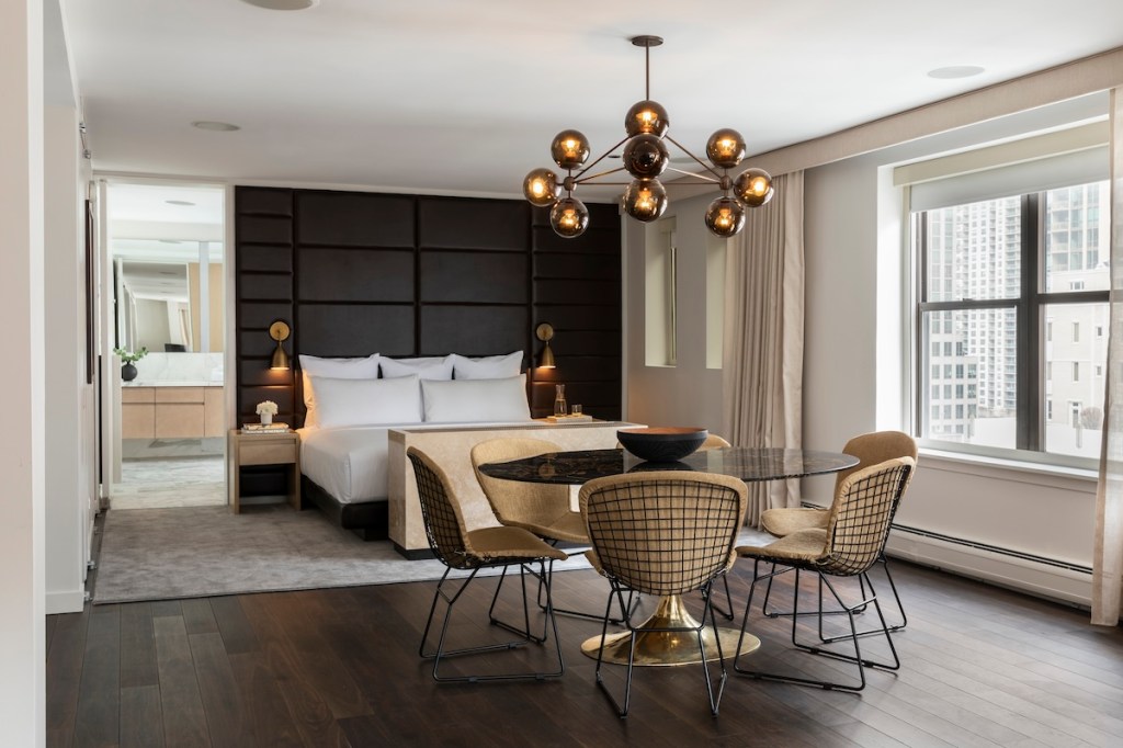 Large hotel suite with round dining table, cream chairs, brass modern chandelier, and large white bed