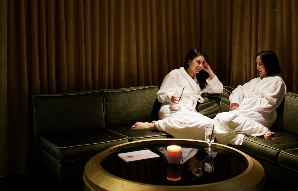 Two women in white robes lounge on a sectional sofa in a dark room