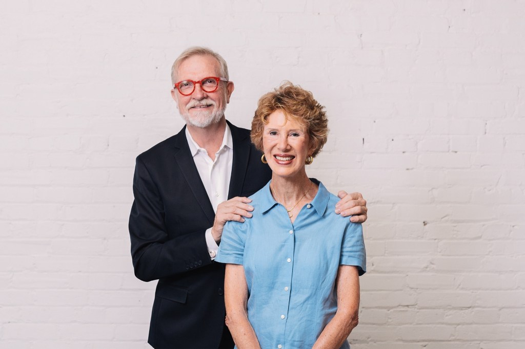 Man in suit coat and red glasses stands with arms around woman in blue shirt