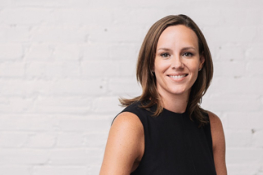 Woman with brown hair in a black top smiles at a camera with a white brick wall behind her