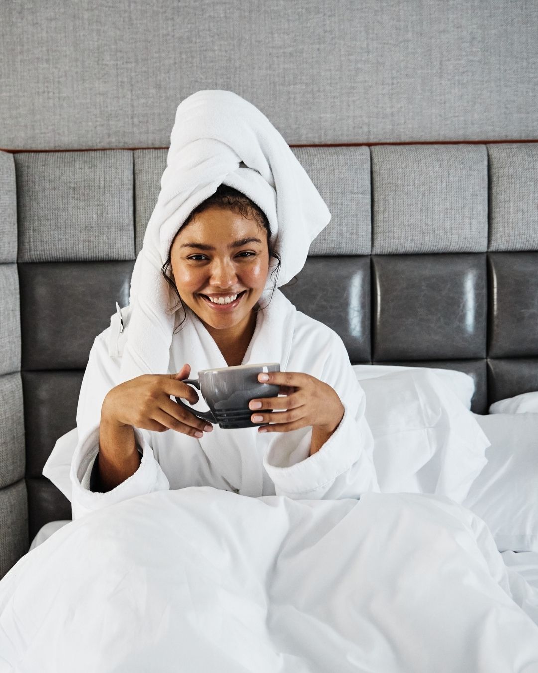 A woman smiling with a mug in hand and hair wrapped in a plush hotel towel