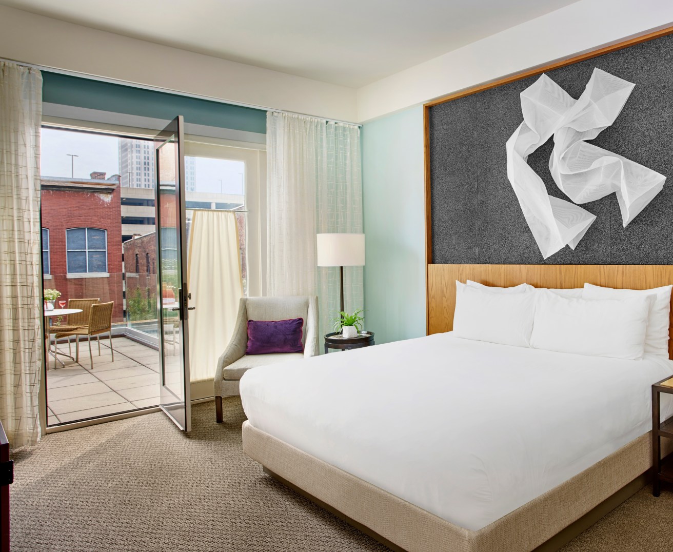 A hotel room with a king bed, artwork above the bed and a door that opens to a large outdoor balcony