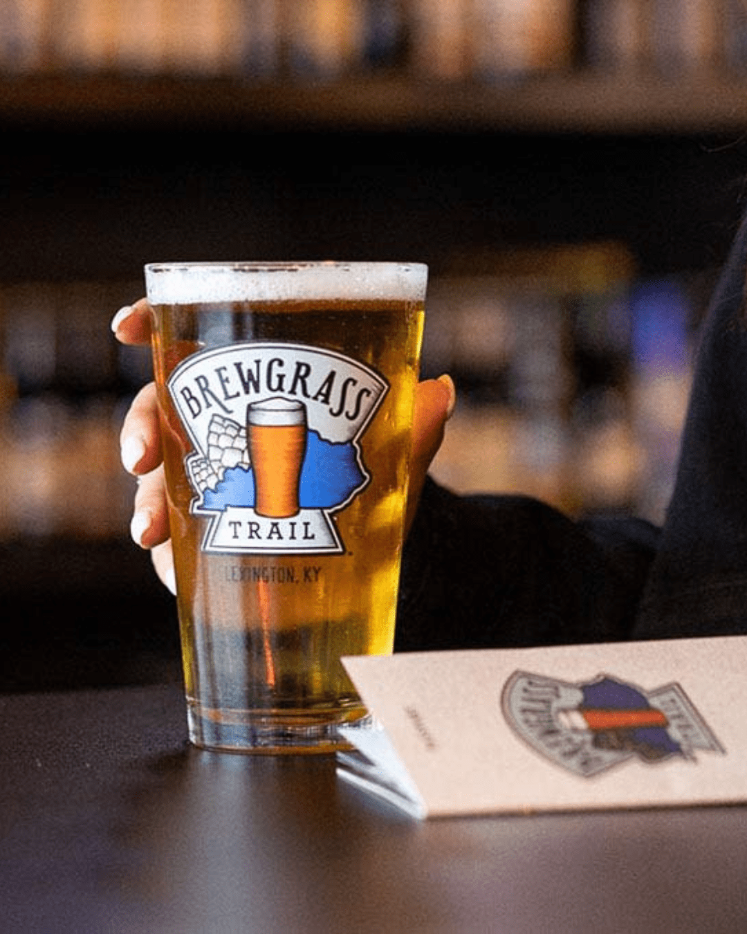 Hand holding beer in glass with Brewgrass Trail logo.