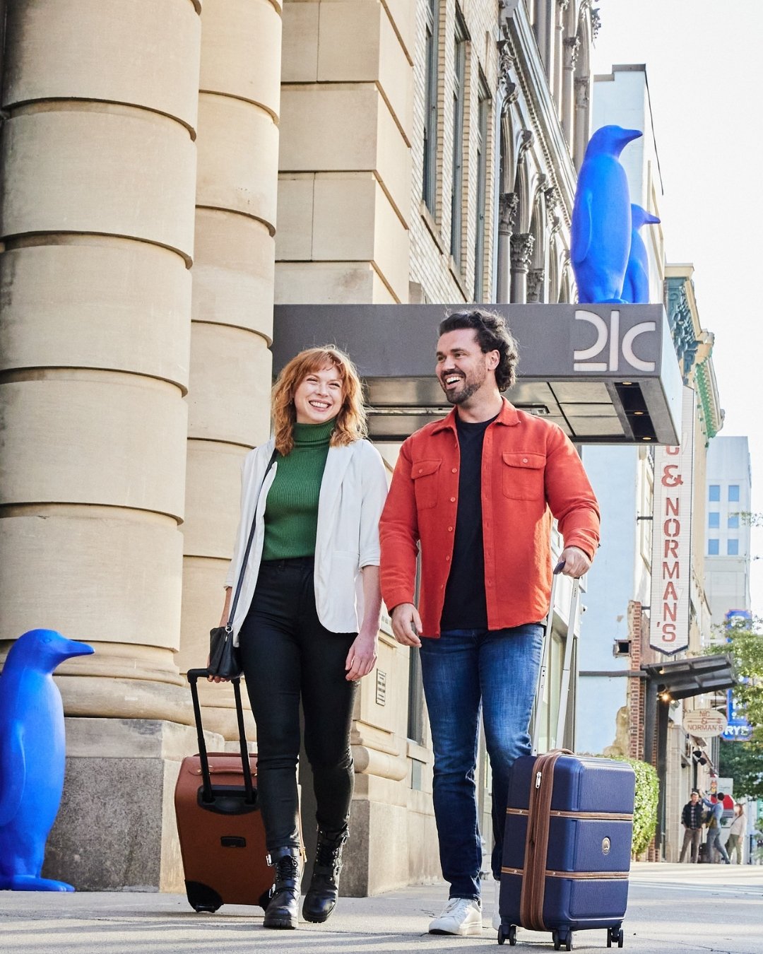 Man and woman roll suitcases hand in hand in front of building with blue penguin statues in the background