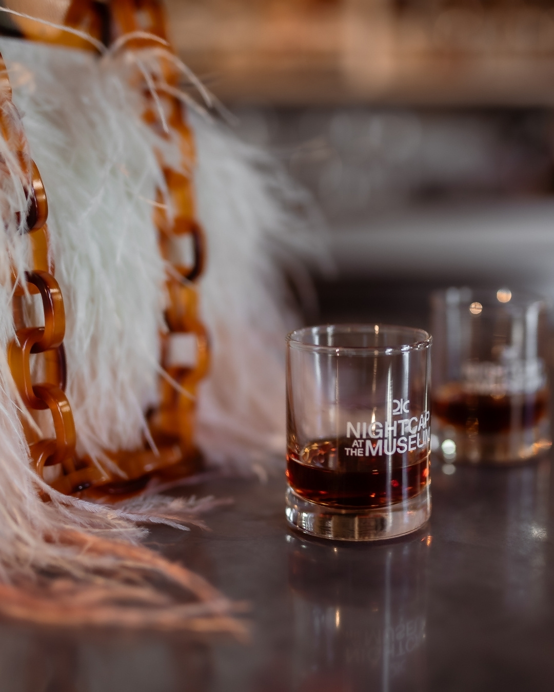 Two small glasses of bourbon on a table with a furry purse nearby
